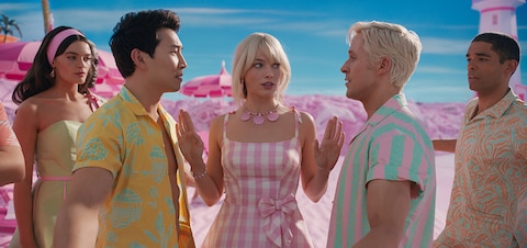 Barbie (Margot Robbie) holds up her hands between two Kens (Simu Liu and Ryan Gosling) to diffuse tension while another Barbie and Ken look on.