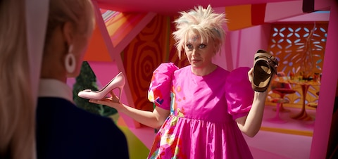 Weird Barbie (Kate McKinnon) holds a pink high heel in her right hand and a brown sandal in her left hand.