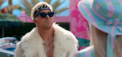 Ken (Ryan Gosling), dressed in a fur coat, black sunglasses, a bandana, and a necklace with a horse pendant, looks at Barbie defiantly.