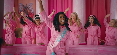 President Barbie (Issa Rae) raises her right arm in triumph as a panel of six other Barbies cheer behind her.