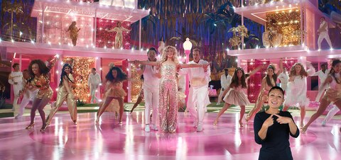 Barbie (Margot Robbie) holds out her arms, about to be lifted up by two Kens in a choreographed dance scene. In the bottom right corner, ASL performer Leila Hanaumi signs "Count on me.”