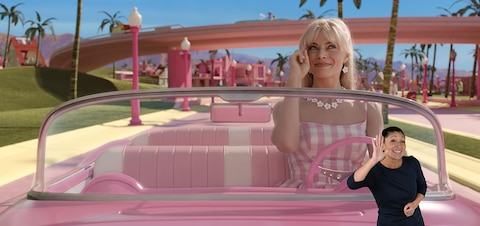 Barbie (Margot Robbie) smiles as she drives her pink convertible through Barbieland. She looks up to the sky and salutes a friendly hello. In the bottom right corner, ASL performer Leila Hanaumi signs “Hello.”