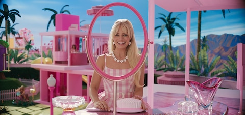 Barbie (Margot Robbie) sits at a vanity table smiling at a mirror with no glass while holding a giant hairbrush.