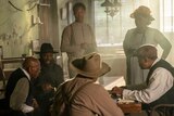 Behind the scenes, Blitz Bazawule speaks to a group of men seated around a gaming table as Corey Hawkins and Danielle Brooks look on.