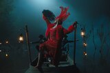 In the foggy night, Shug — wearing a red beaded dress, red fur coat, and red feather headpiece — strikes a dramatic pose on the bow of a rowboat while holding a black feather fan. Two men in suits behind her reach out to the side.