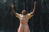 Young Celie smiles as she stands in the rain, her face and arms raised toward the sky.