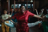 Celie smiles jubilantly during a choreographed dance — Sofia to her left and Shug to her right — while a male dancer behind holds out her arms. A group of dancers behind them smile with their arms straight up.