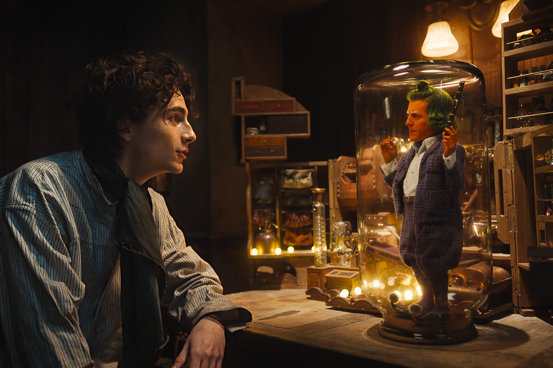 Willy Wonka sits at a desk talking to Oompa Loompa. Oompa Loompa, a small man with orange skin and green hair, stands inside a glass jar on the desktop.