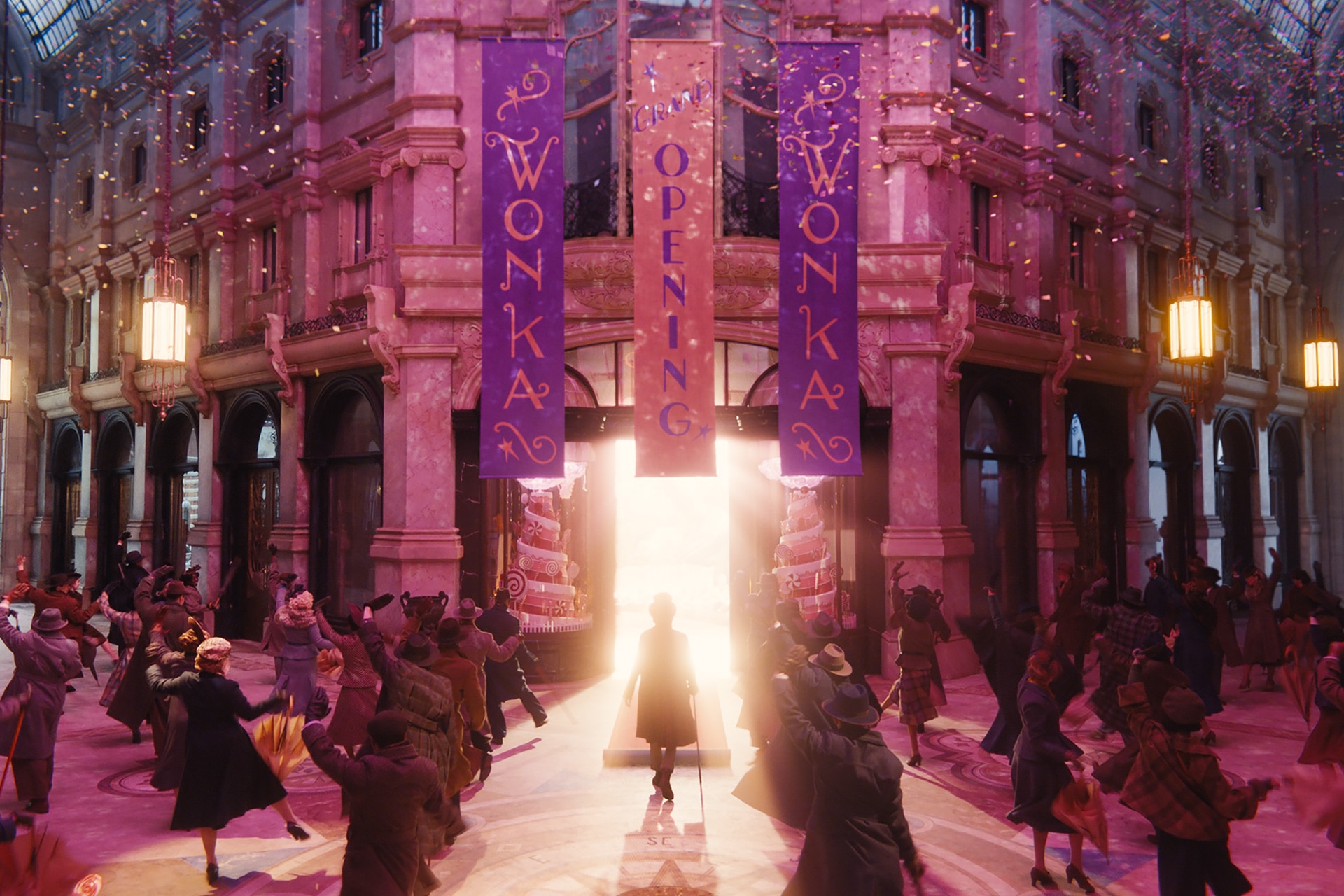 A crowd of people is gathered outside a large building decorated with three purple and gold banners that read "Wonka," "Grand Opening," "Wonka." A bright light glows from the entrance and illuminates Willy Wonka. Confetti is in the air.
