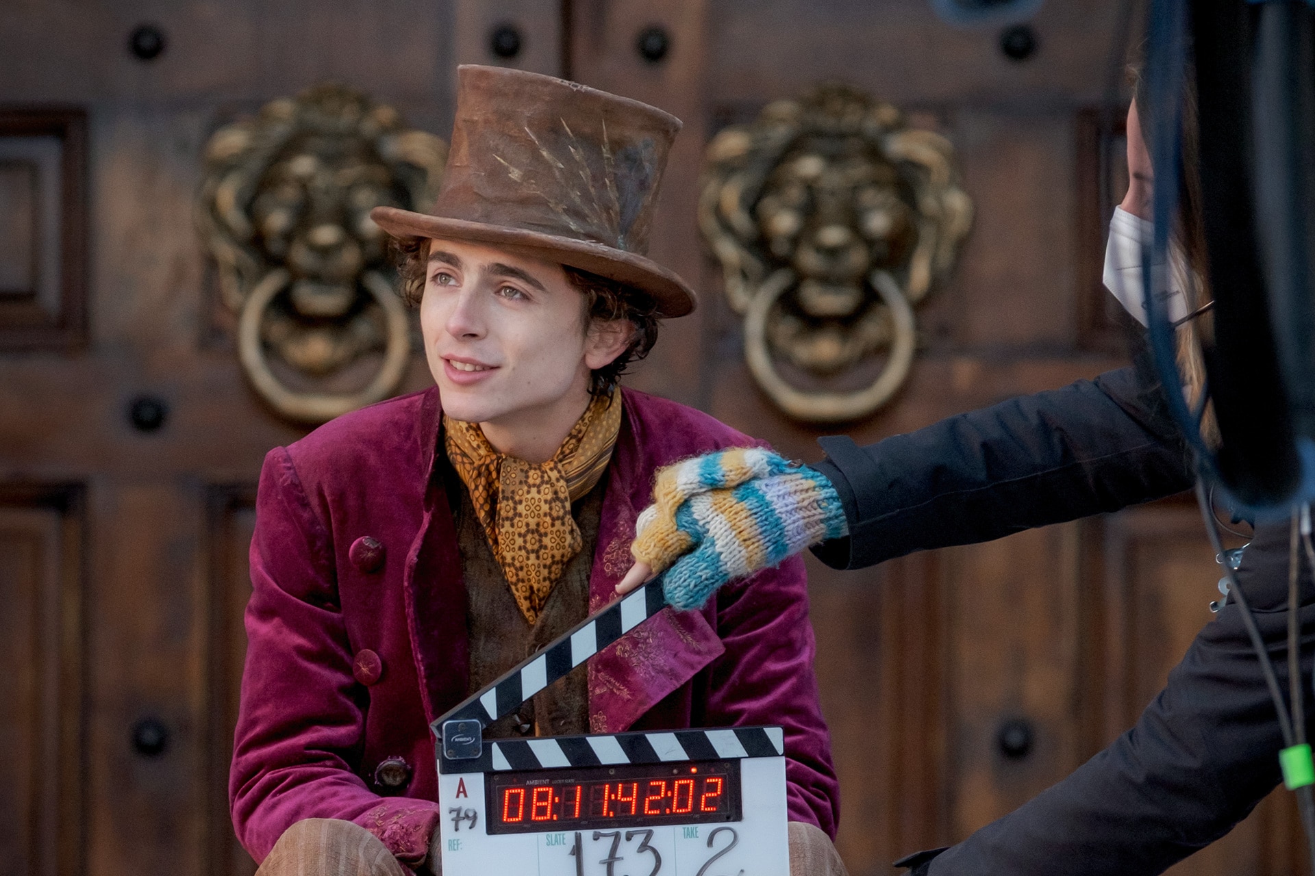 Behind the scenes, Timothée Chalamet, in costume as Willy Wonka, sits on stone steps in front of large wood doors. A boom mic is overhead and a crewperson holds a clapper board that reads, "Slate: 173, Take: 2, Wonka, 3.11.21, Director: Paul King." 