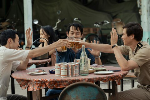 Three smiling men with raised left hands, including The Captain in a military uniform, clink glasses of beer while seated at a round table covered in a plaid tablecloth, cans of beer, packs of cigarettes, and empty plates.
