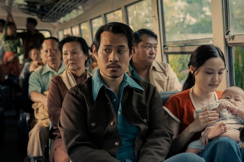 Bon is seated next to a young woman holding a baby on a crowded bus.