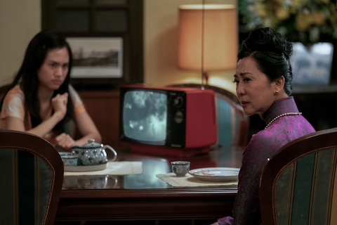 Madame sits at a dining table with a young woman. There is a blue and white tea set and a red TV on the tabletop.