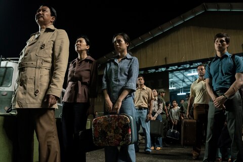 A crowd of people, including The General, Madame, and The Captain, stand outside an airplane hangar at night, holding suitcases.