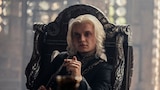 King Aegon II Targaryen sits in an elaborately carved high-back chair, holding a dagger. 