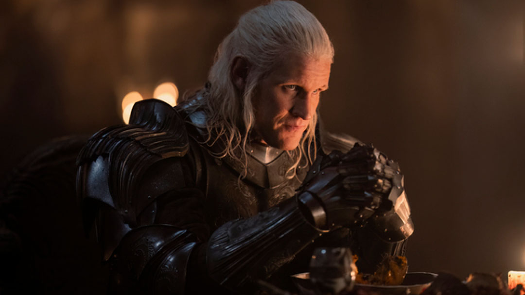 Prince Daemon Targaryen sits at a dining table in a suit of armor, his hands clasped together. 
