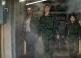Trapper, Ilene Andrews, and Jia walk down an industrial hallway wearing green jumpsuits.