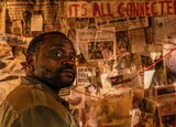 Bernie Hayes stands in front of a board covered in newspaper clippings and a red string. A handwritten piece of paper at the top reads “It’s All Connected...”