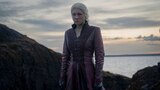 Queen Rhaenyra Targaryen stands on a rock formation with a body of water behind her.
