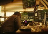 ASL performer Otis Jones signs “provides” in front of a scene from the movie that shows Bernie Hayes sitting at a cluttered desk, looking at threelarge computer monitors.