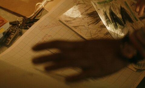 ASL performer Otis Jones signs “Home” in front of a scene from the movie that shows hands reaching for a large piece of graph paper titled “Seismic Shift” and small pieces of paper covered in triangular scribbles.