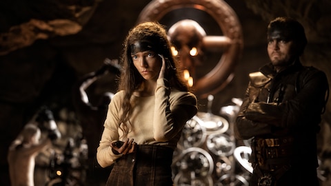 Furiosa, with black paint covering her forehead, stands with her left hand on her temple. Praetorian Jack and an altar of steering wheels stand behind her.