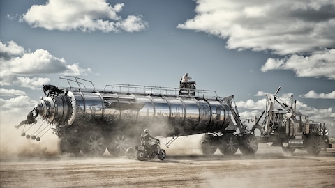 The War Rig, a gleaming silver semitruck embossed with a battle scene, races across the Wasteland. War Boys try to defend the truck as two members of the Biker Horde climb up the back and another rides alongside on a motorcycle. 