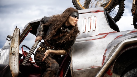 Furiosa exits a silver vehicle while holding a long revolver in her right hand. 