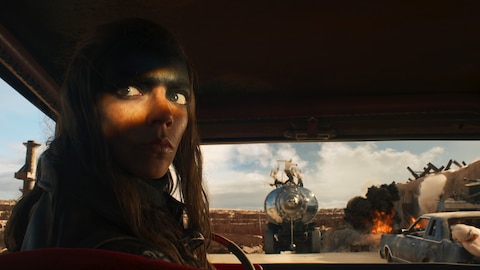Close-up of Furiosa looking over her shoulder as she sits behind the wheel of a vehicle. In front of the vehicle is the War Rig with a group of War Boys standing on top. A fire emitting thick, black smoke burns to the right. 