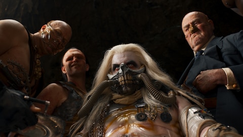 Rictus Erectus, Scabrous Scrotus, The Immortan Joe, and The People Eater stand in a semicircle and look down at an object being presented to them. 