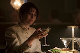 Sofia Falcone sits at a dining table and raises a martini glass in a toast