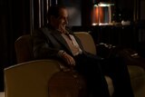 Oz Cobb sits in an armchair and laughs at something out of frame.
