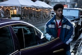 Victor Aguilar stands outside a Maserati sedan and holds the rear passenger door open.