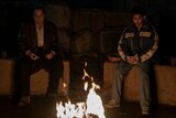Oz Cobb and Victor Aguilar sit on cement blocks in front of a small fire and hold sandwiches.