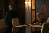 Milo Grapa stands next to and Johnny Vitti sits at a conference table. They both look at something out of frame.