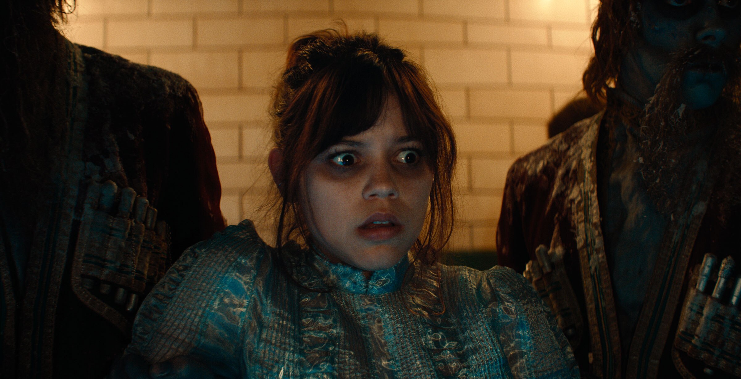 Two dark figures hold Astrid Deetz by the arms as she looks at something out of frame with concern.
