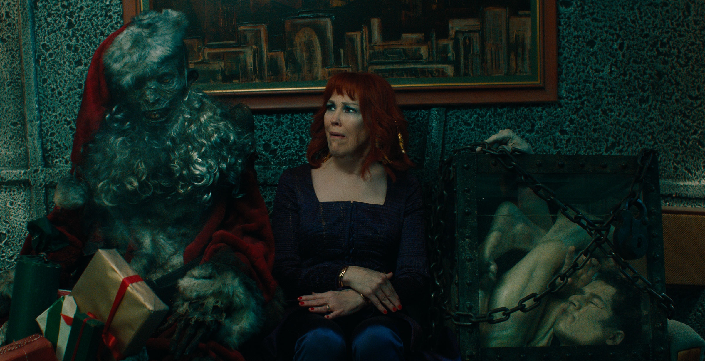 Delia Deetz looks horrified as she sits between a dirty skeletal Santa Claus and a man contorted inside a chained, glass box.