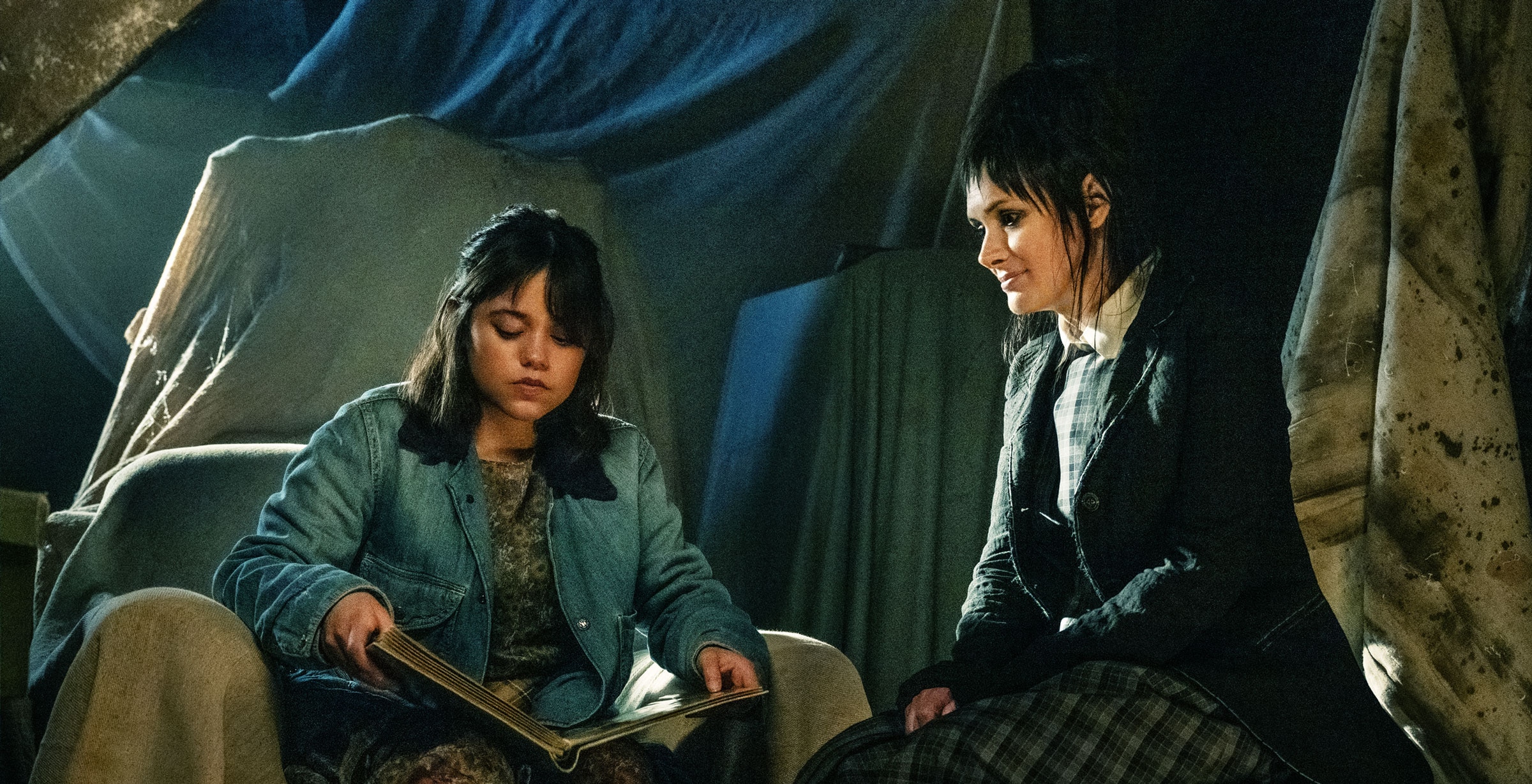 Astrid Deetz reads from a large book while seated next to Lydia Deetz in a room where the furniture is covered in sheets.
