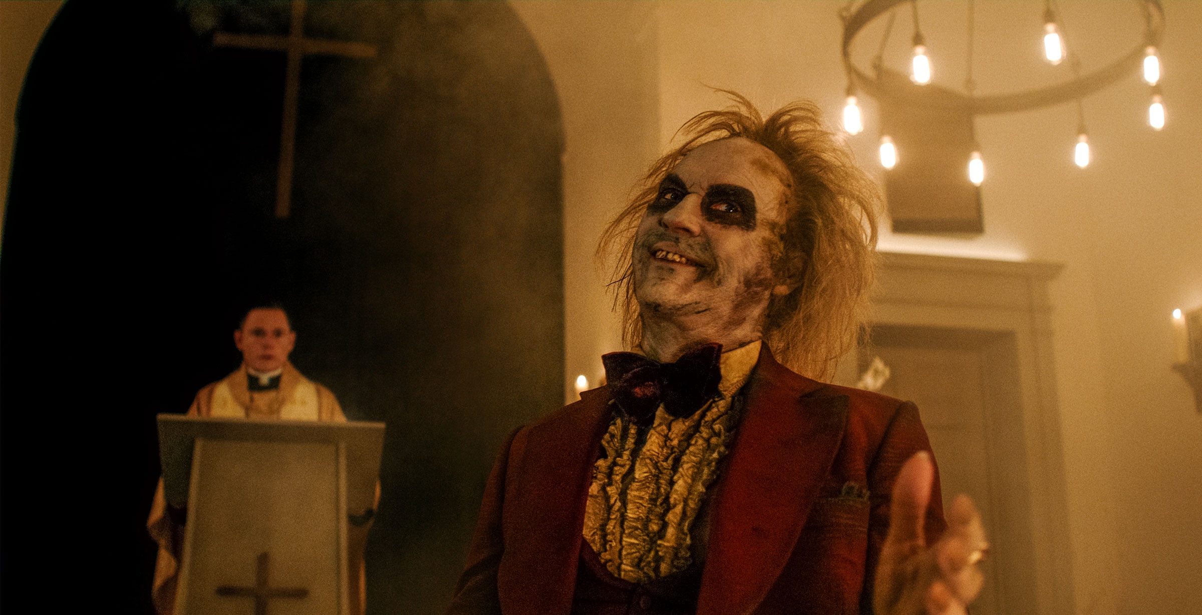 Beetlejuice is dressed in a decaying red tuxedo and stands in front of a priest who is at an altar. He smiles wryly as he gestures at something out of frame.