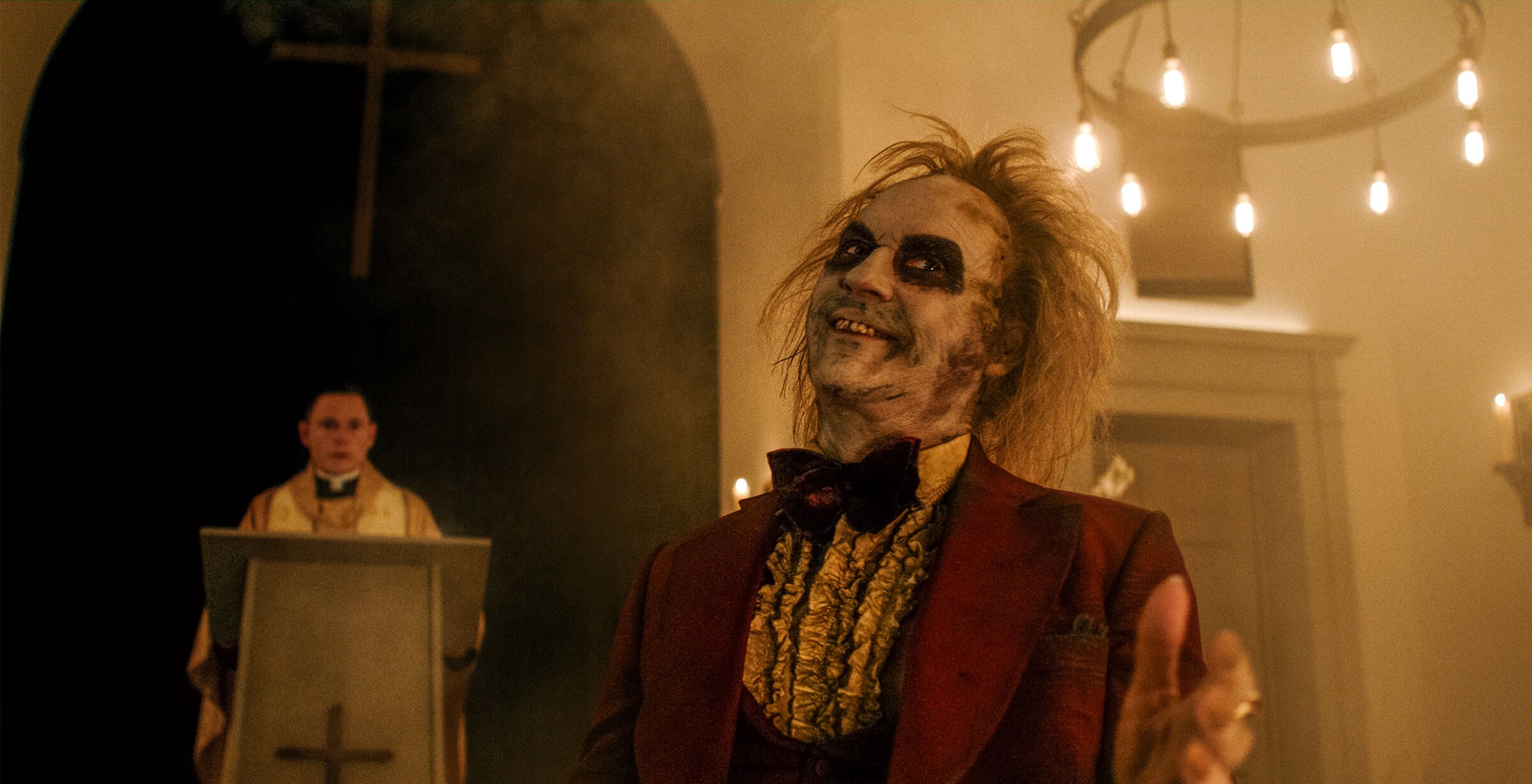 Beetlejuice is dressed in a decaying red tuxedo and stands in front of a priest who is at an altar. He smiles wryly as he gestures at something out of frame.