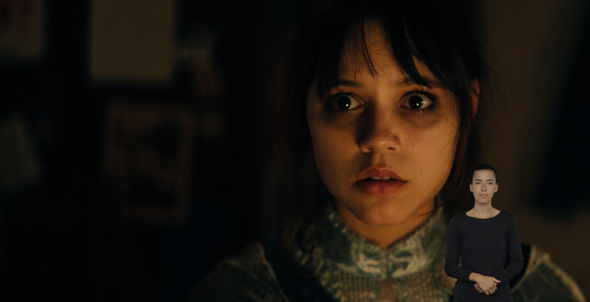 Astrid Deetz looks at something out of frame. Her eyes have dark circles underneath them. ASL performer Sophia Morales stands in the bottom right with her hands clasped.