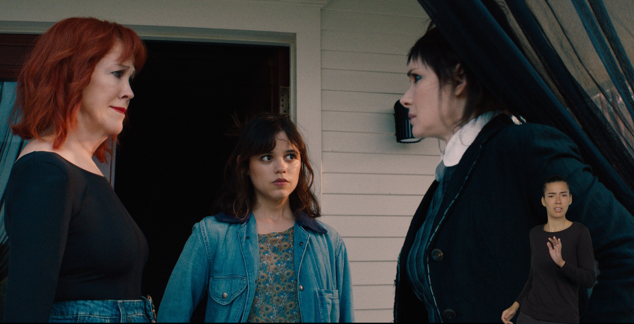 Delia Deetz and Astrid Deetz look at Lydia Deetz who is holding open a sheer, black curtain. ASL performer Sophia Morales stands in the bottom right and signs “wait.”