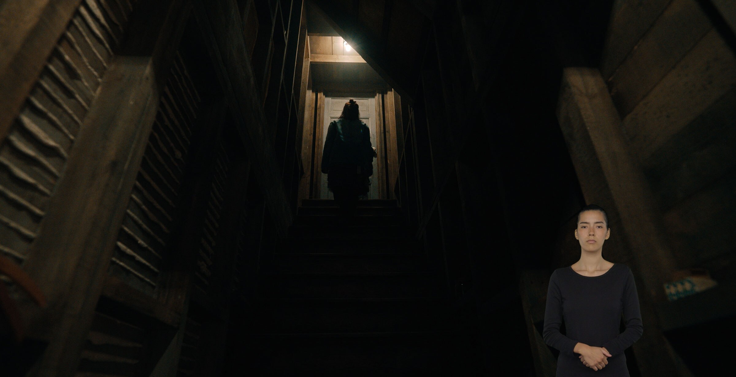 Astrid Deetz stands at the top of a staircase in front of a closed door. It’s dark except for a single lightbulb shining above her. ASL performer Sophia Morales stands in the bottom right with her hands clasped.