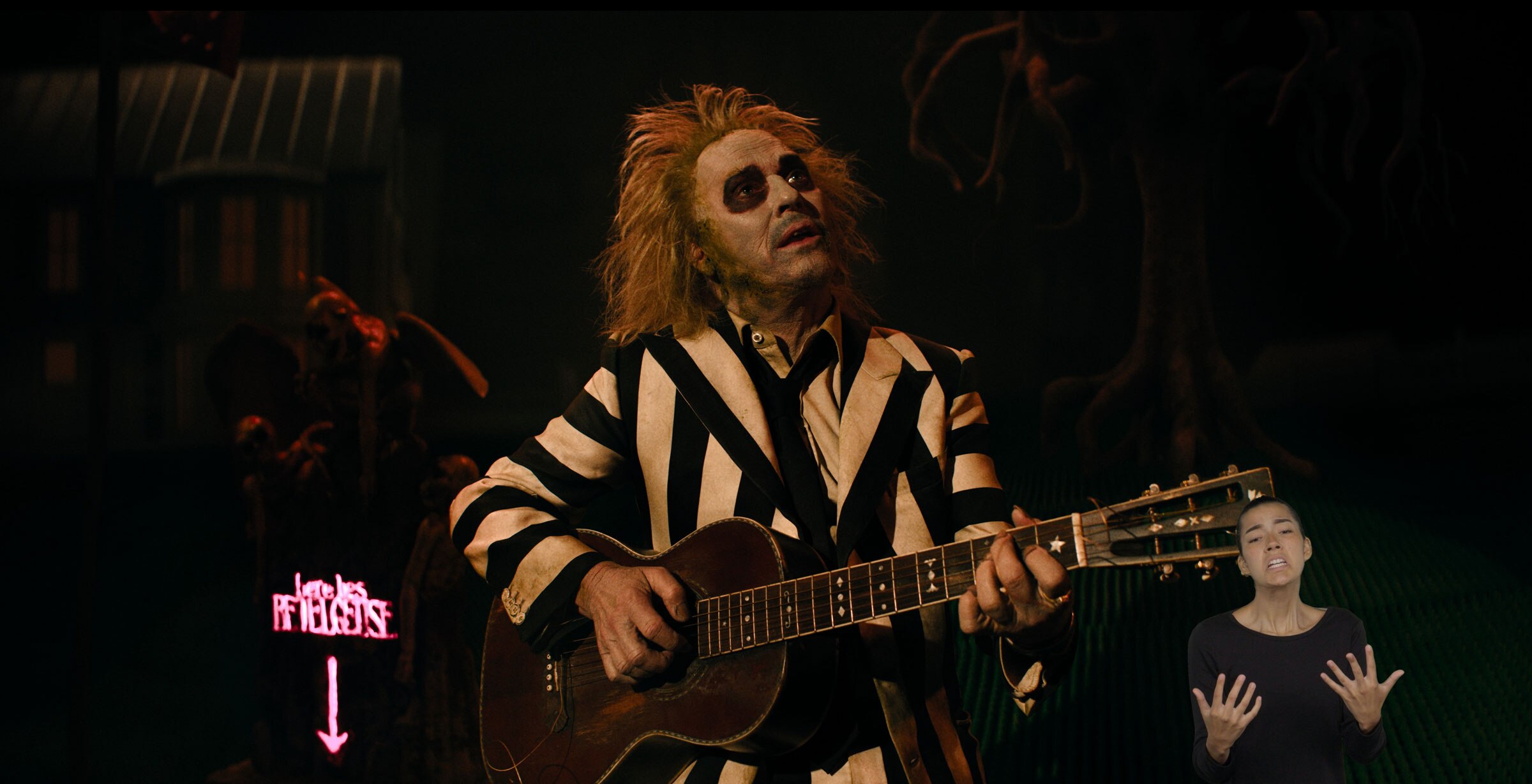 Beetlejuice plays an acoustic guitar and looks at something out of frame. A tombstone behind him reads, “Here lies Betelgeuse.” ASL performer Sophia Morales stands in the bottom right and signs “waiting.”