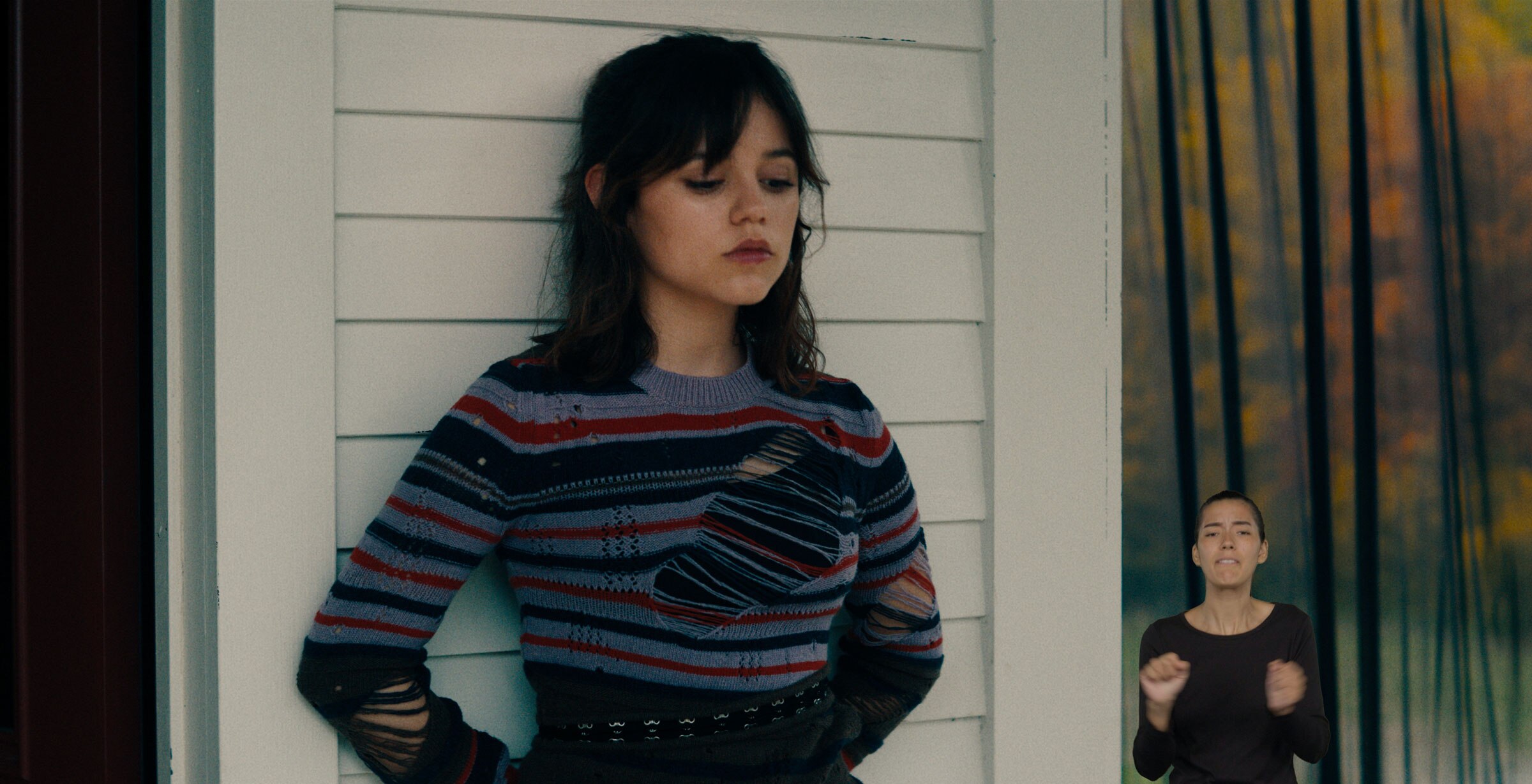 Astrid Deetz leans against a white clapboard building with her eyes downcast. ASL performer Sophia Morales stands in the bottom right and signs “safe.”