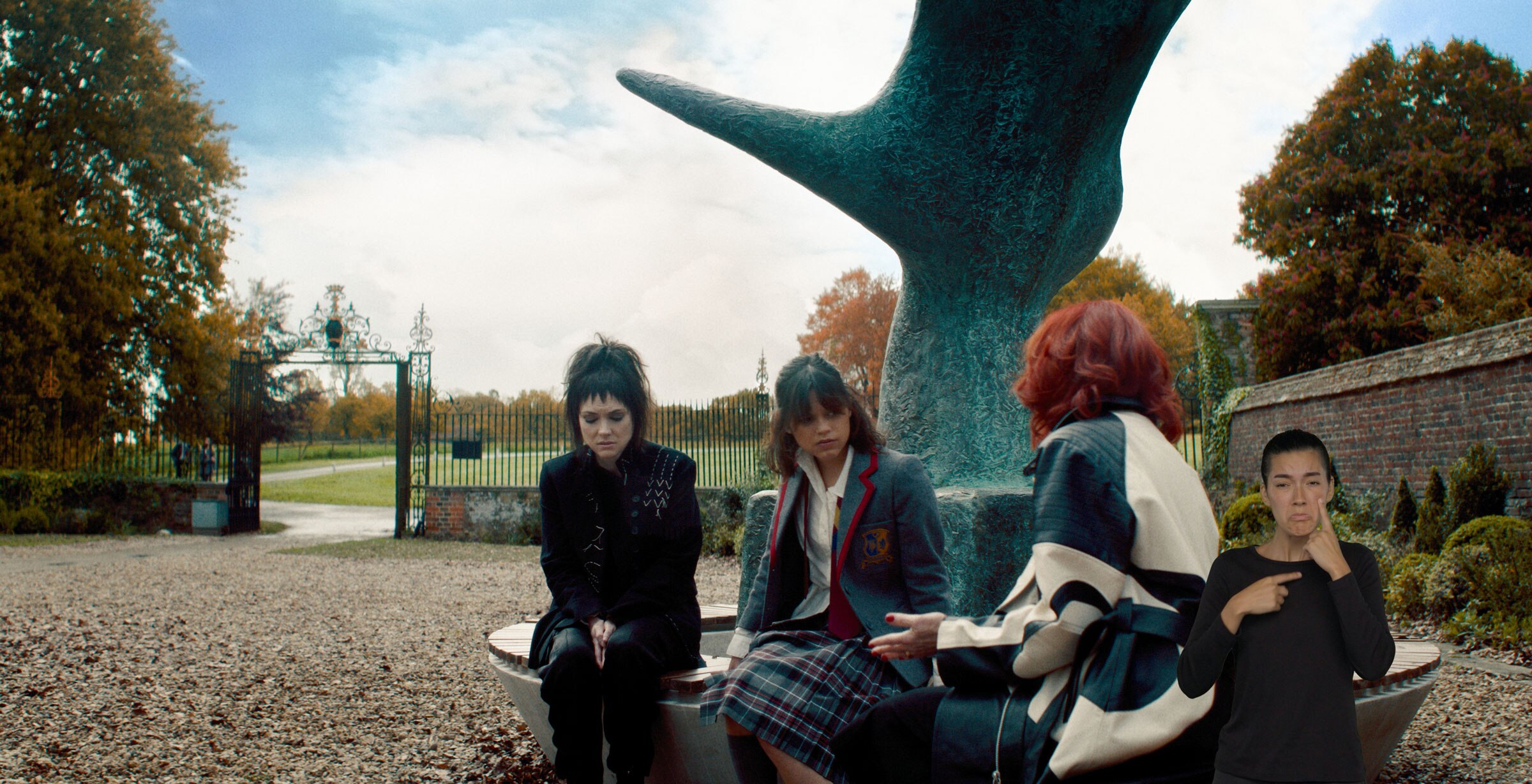 Lydia Deetz, Astrid Deetz, and Delia Deetz have a conversation as they sit on a bench around a large sculpture. ASL performer Sophia Morales stands in the bottom right and signs “tears.”