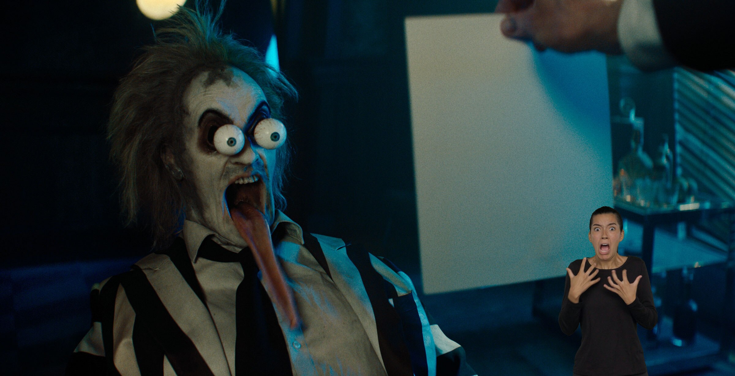 Beetlejuice cartoonishly screams with large bulging eyes, mouth wide open, and a ridiculously long tongue hanging out. ASL performer Sophia Morales stands in the bottom right and signs “screaming.”