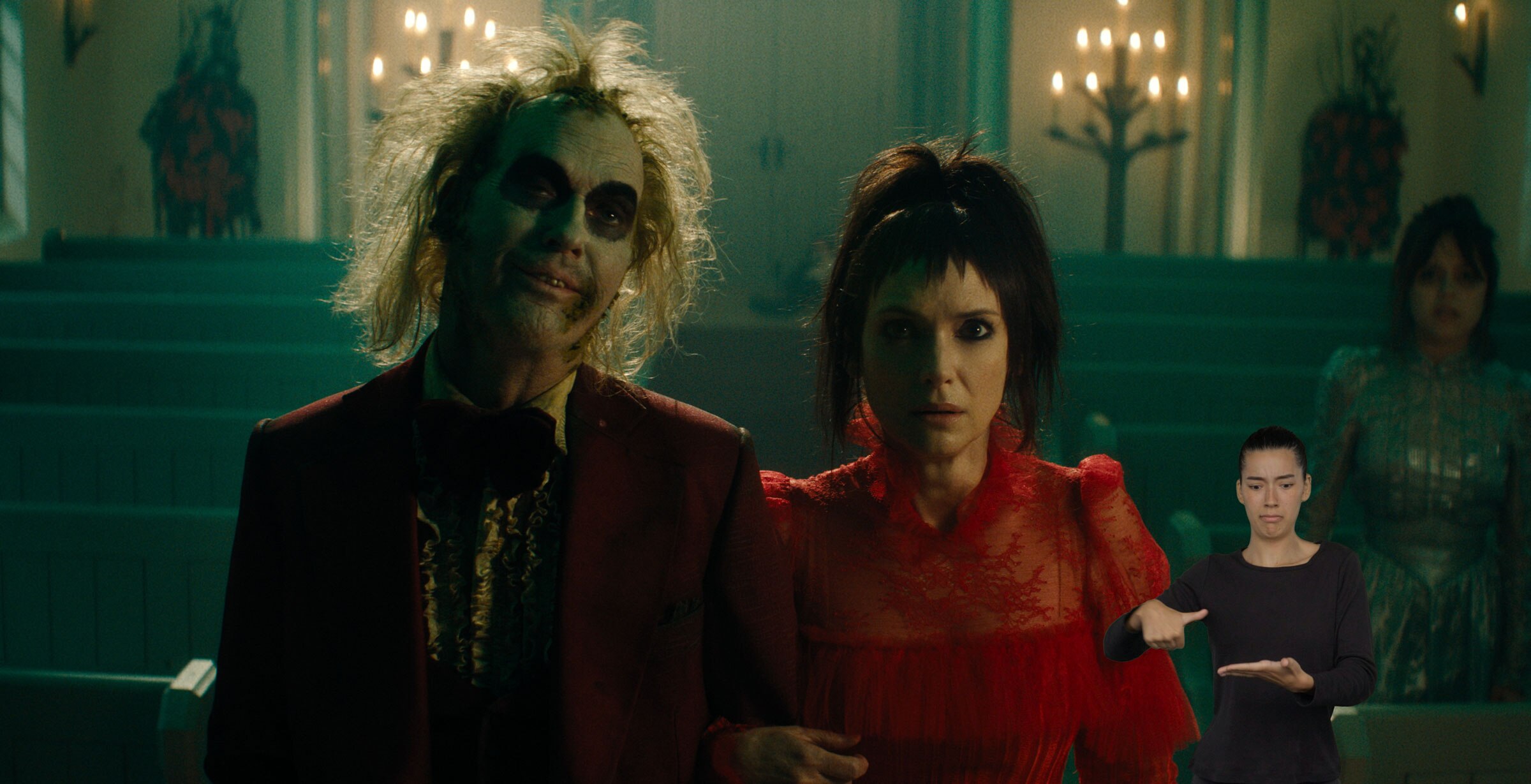 Beetlejuice and Lydia Deetz stand arm-in-arm inside of a candle-lit church as Astrid Deetz looks on in the background. Beetlejuice wears a shabby red suit, and Lydia wears a formal red dress. ASL performer Sophia Morales stands in the bottom right and signs “iron.”