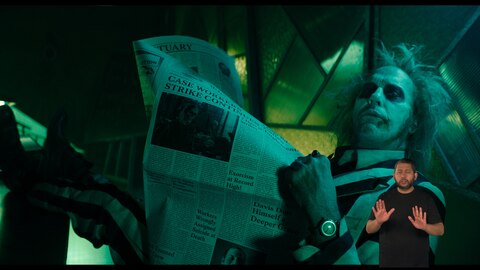 Beetlejuice está relaxado, com as pernas apoiadas e lendo um jornal com expressão desinteressada. No canto inferior direito, o intérprete de Libras, Andrey Baptista, sinaliza "esperar".