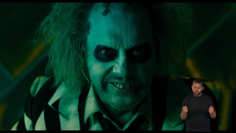 Beetlejuice olha fixamente para a câmera e sorri de forma maliciosa. O intérprete de Libras, Andrey Baptista, está no canto inferior direito e sinaliza "agarrar".